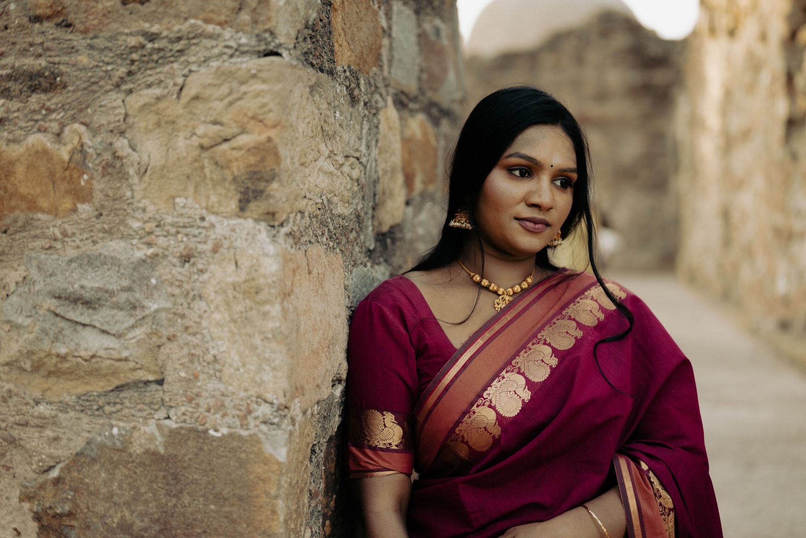 Elegant portrait of a woman in a red sari leaning against a stone wall.